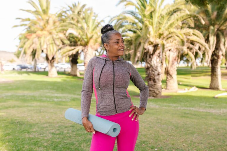 Senior woman smiling with yoga mat in a sunny park, embodying active lifestyle and wellness.