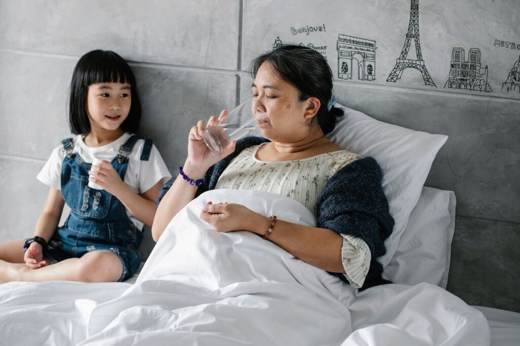 Upset sick aged Asian woman drinking water after taking medicine while lying in bed near cute little granddaughter