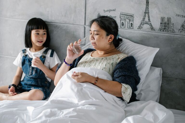 Upset sick aged Asian woman drinking water after taking medicine while lying in bed near cute little granddaughter