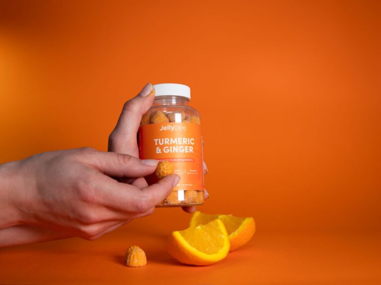 Hand holding Jellybee Turmeric & Ginger gummies with orange slices on vibrant orange background.